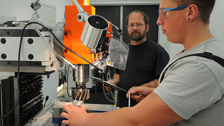 Junger Mann arbeitet an einer Werkzeugmaschine, daneben steht sein Ausbilder.