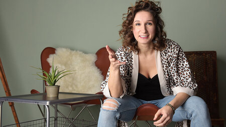 A woman wearing jeans and a short-sleeved cardigan sits on a couch.