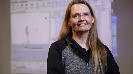 A woman with long hair and glasses stands on front of a blackboard.