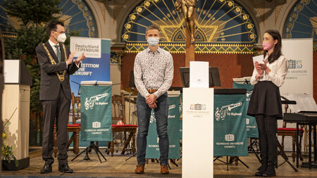 Ein Mann mit Maske und Anzuge applaudiert einem Man mit Maske, der eine Urkunde hält.