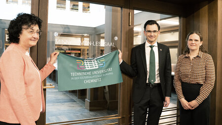 Ein Mann und eine Frau halten ein Textilbanner unter einen Schriftzug an einer Glastür. 
