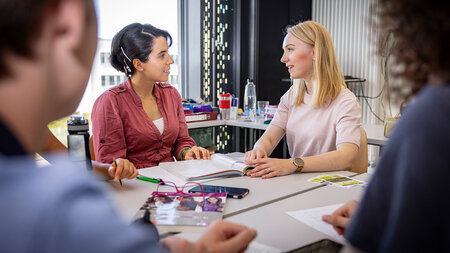 Zwei junge Frauen sitzen an einem Tisch und sprechen miteinander.
